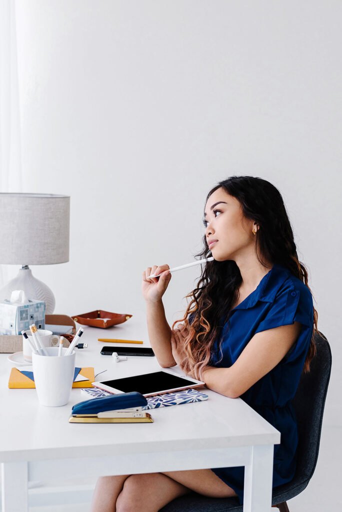 A woman with a navy shirt and dark hair is sitting at a white desk looking deep in thought. She is holding a pencil to her chin and there is a container of more pencils nearby and her desk has a notebook and tablet in front of her.