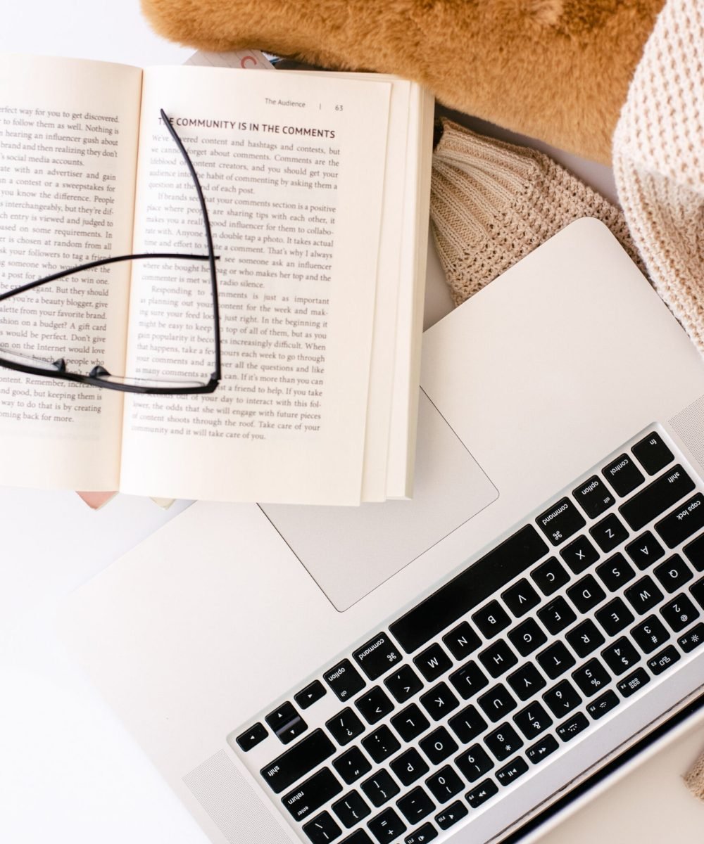 An image of a tan pillow, open book with glasses laying on it and an open laptop