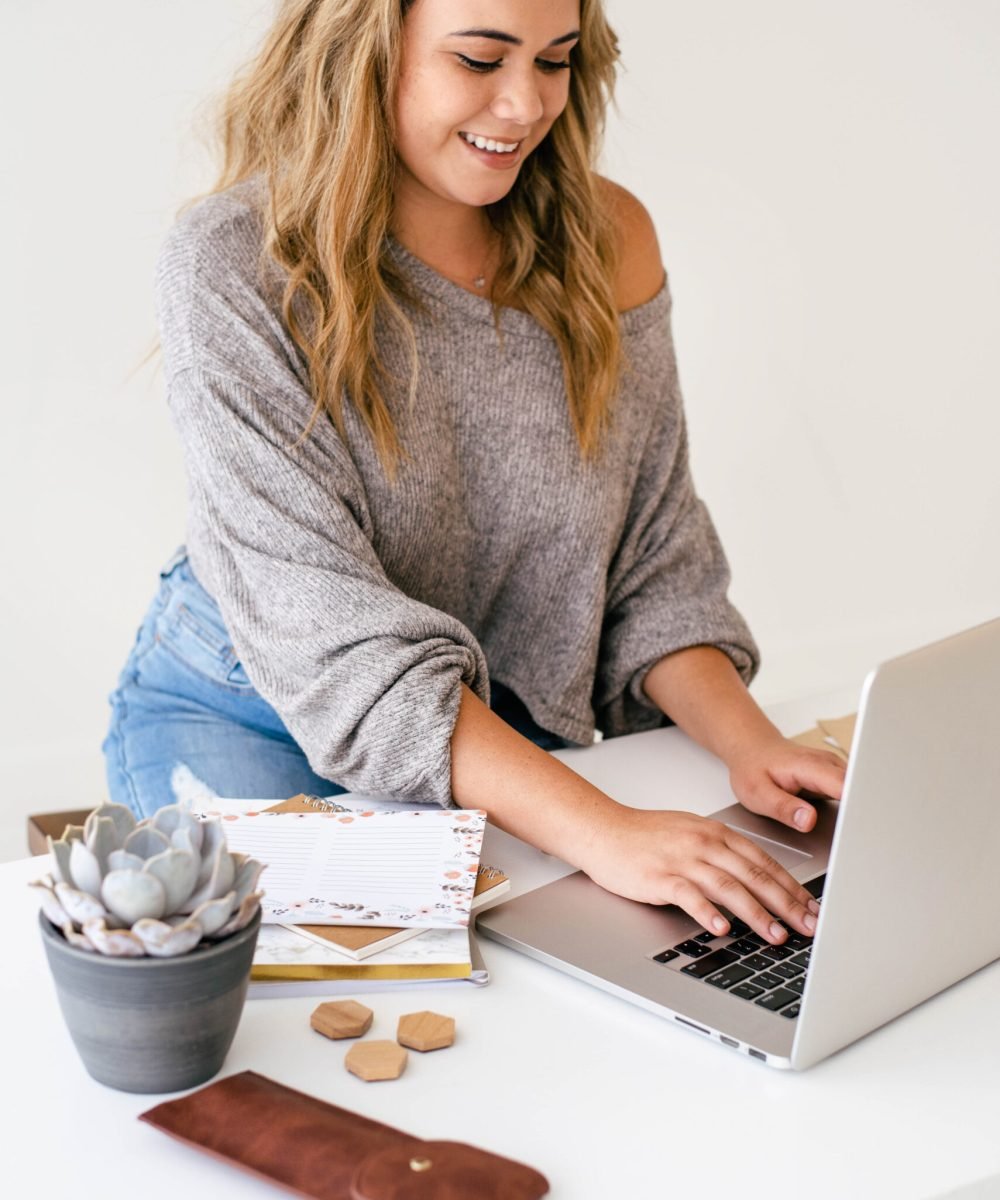 A woman with wavy hair wearing a grey sweatshirt and jeans typing on a laptop. There is a container and a brown leather sleeve on the white desk next to her.