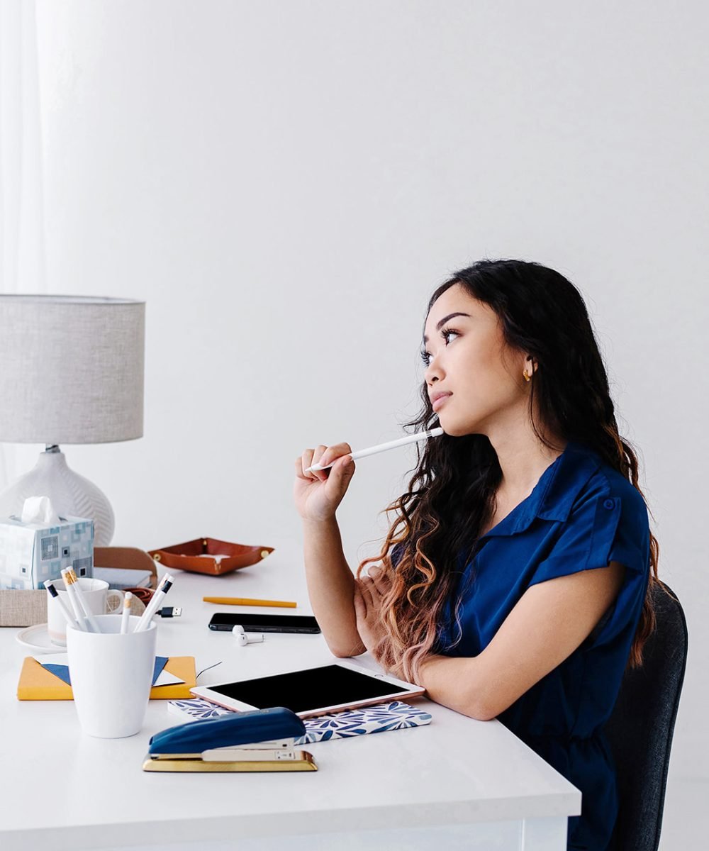 A woman with a navy shirt and dark hair is sitting at a white desk looking deep in thought. She is holding a pencil to her chin and there is a container of more pencils nearby and her desk has a notebook and tablet in front of her.